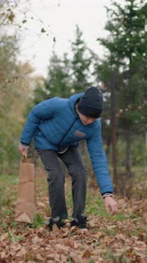 Boy Collecting Leaves in Paper Bag with Fence and Building in Background