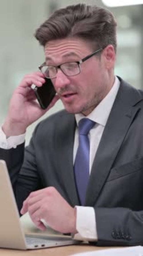 Middle Aged Businessman with Laptop Talking on Smartphone in Office, vertical video