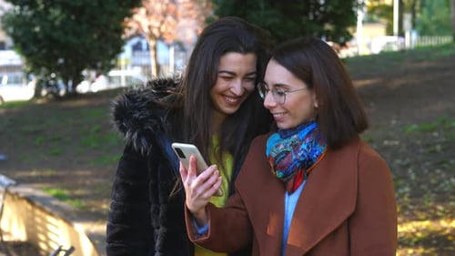 app, technology - young women at park laugh looking at photos on phone