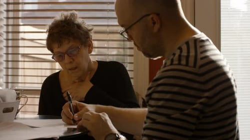 Madre mayor firmando documentos con la ayuda de su hijo cuidador en casa en una tarde de primavera (Medium Shot)
