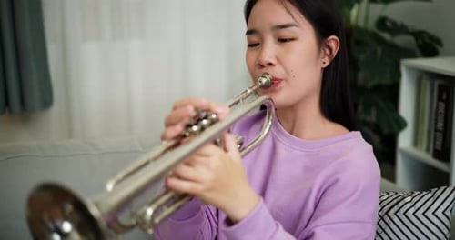 A young woman practices playing the trumpet while sitting on a sofa in a cozy living room.