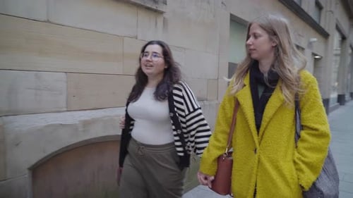 Two young lesbians walking hand in hand through part of a city and chatting