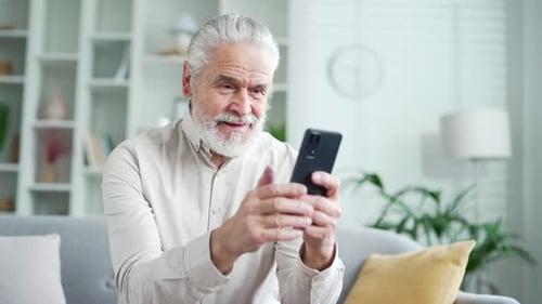 Happy elderly senior man with gray hair received great news on smartphone while sitting on sofa