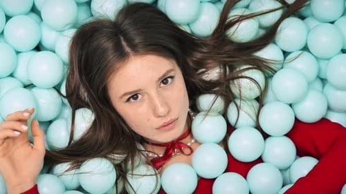 Young woman in a red dress lying and posing in a pile of plastic blue balls in a studio