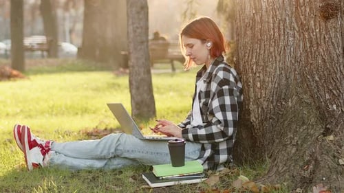 Young Attractive Girl with Red Hair Wearing Casual Clothes Sitting Under the Tree in a City Park on