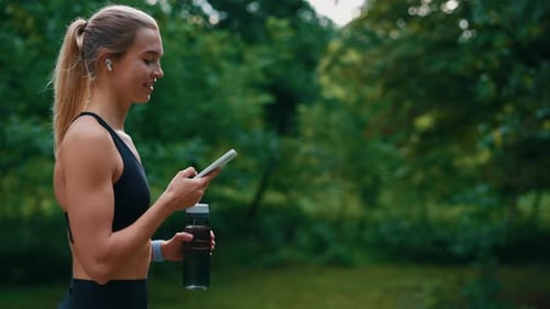 Side View of the Sporty Woman Walking Holding Sport Bottle Using Scrolling Phone and Listening to