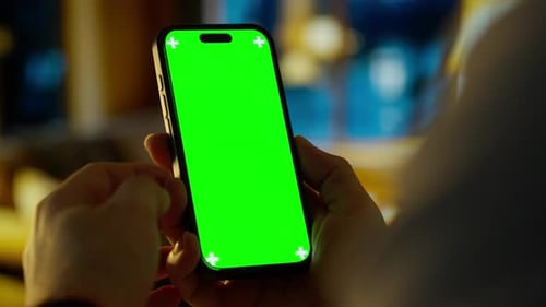 Person Holding Modern Smartphone with Bright Green Screen Mockup in Cozy Indoor Home Environment