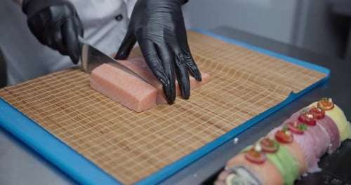 Un chef portant des gants en nitrile noir prépare des sushis en tranchant le saumon avec un couteau sur une planche à découper