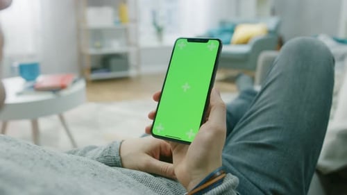 Man at Home Lying on a Couch using Smartphone with Green Mock-up Screen, Doing Swiping, Scrolling G