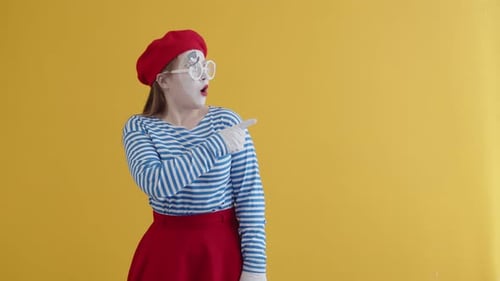 Pantomime Frau in Make-up, rote Baskenmütze, gestreiftes Hemd, roter Rock, streckt ihren Finger zur Seite und ist sehr