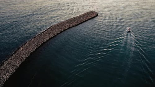 Boat glides past rocky jetty on calm water during sunset