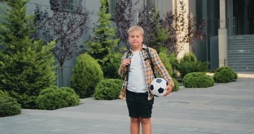 Portrait of confident blonde boy posing with footbal ball outdoors modern school building background