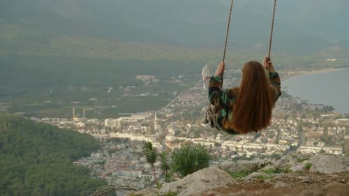 With a view of the sea and the city, a young woman swings on swings over the cliff from a high mount