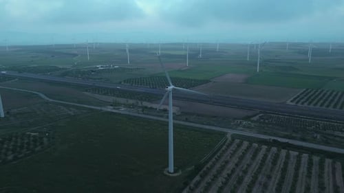 Drone orbit around wind turbine at dawn over structured farmland and highway
