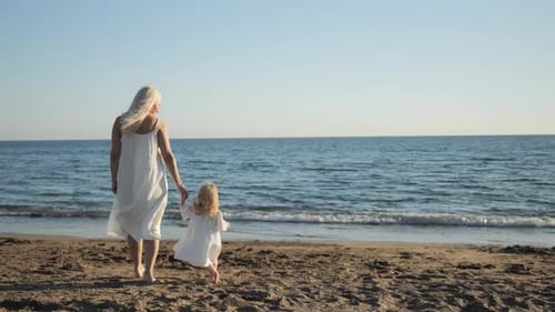 Mom and Daughter in White Dresses Walking on the Sea Beach at Sunset Mom and Daughter in White Dresses Walking on the Sea Beach at Sunset