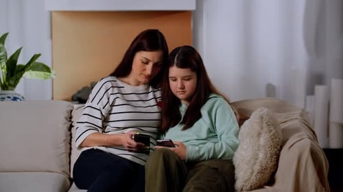 The Girl and Her Mother are Sitting on the Sofa in the Living Room with Phones in Their Hands Mom