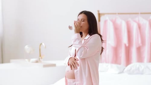 Schwangere Frau im rosa Pyjama lächelt, während sie in einem hellen Raum Wasser trinkt