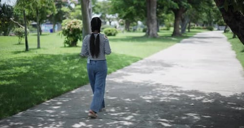 Young woman walks leisurely along a concrete path in a quiet open area of a park.