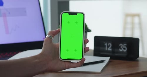 Female Hand Holds Smartphone with Green Screen on Background of Workspace in Bright Room Concept