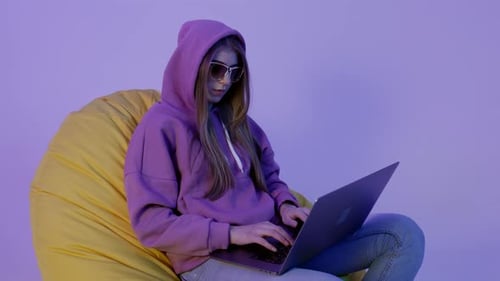 Young Woman Sitting on Pouf and Using Notebook Isolated on Violet Color Background Girl in Hoodie
