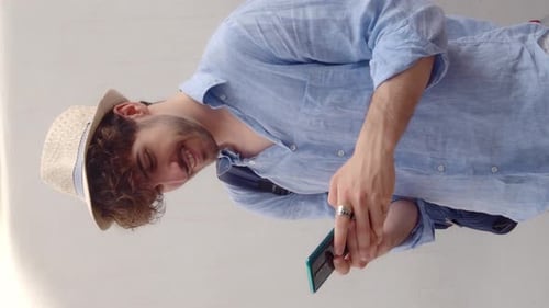Vertical videoYoung Man in Hat Chatting with Smartphone in a Seaside Town During Vacation