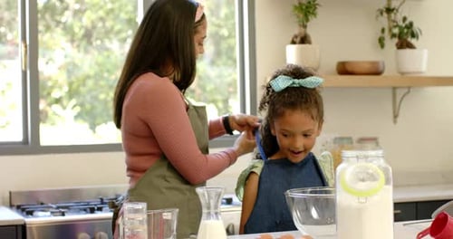 Madre trenzando los rizos de su hija en la isla de la cocina, cogiendo un peine azul y peinando el pelo para la escuela