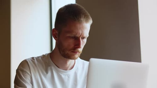 Ein konzentrierter Mann in einem weißen T-Shirt schaut auf den Bildschirm seines Laptops Angespannter Mann, der an einem Laptop arbeitet
