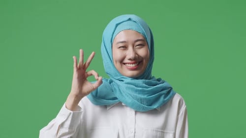 Nahaufnahme einer asiatischen muslimischen Frau, die lächelt und auf dem grünen Bildschirm eine Okay-Geste vor der Kamera zeigt