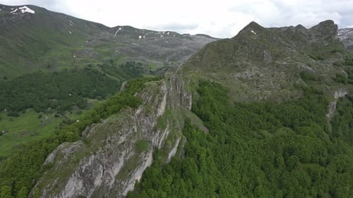 Flying over beautiful green mountains. In the background, stone cliffs with snow are visible.