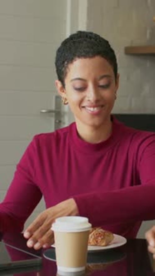 Vertical video: Responding to colleague cue, woman in maroon top tapping tablet at table in kitchen