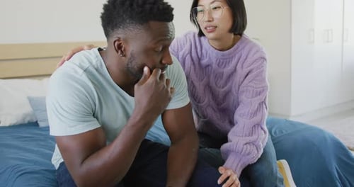 Sad diverse couple sitting on couch and talking in bedroom