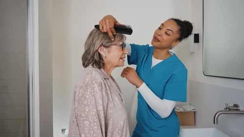Friendly caregiver brushing senior woman's hair in bathroom