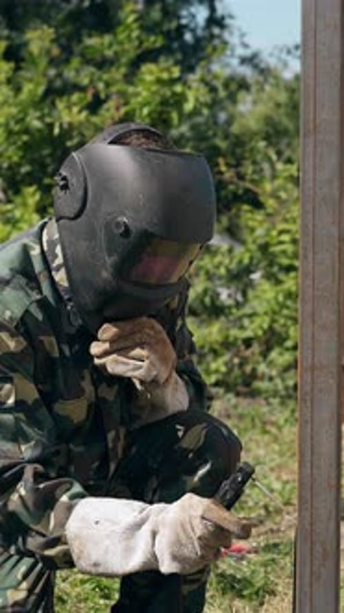 Un reparador de ropa de trabajo suelda la carcasa de una cerca en una casa de campo