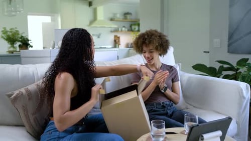 Femmes afro-américaines sur un canapé plaçant une boîte, découpant du ruban adhésif, révélant un objet à bulles pour inspection