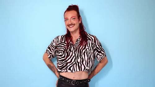 Non-binary person with red hair and mustache smiling on blue background
