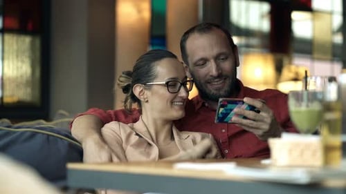 Happy Couple Watching Something On Smartphone Sitting In Cafe