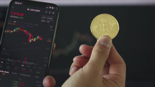 A Closeup of a Hand Holding a Bitcoin Coin and a Smartphone with Stock Charts Stock Reports in the