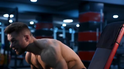 Shirtless bodybuilder in gym. Young guy with muscular body is lifting dumbbells.