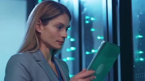 A Woman in a Suit Works Intently on a Tablet in a Server Room