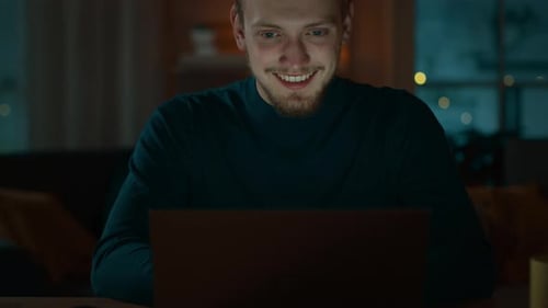 Handsome Smiling Man Works on a Laptop while Sitting at His Desk at Home. Portrait of a Young Freel