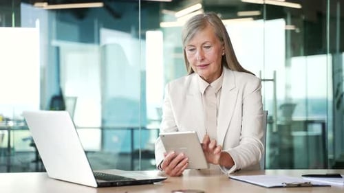 Confident senior gray haired businesswoman is using digital tablet sitting at workplace in business