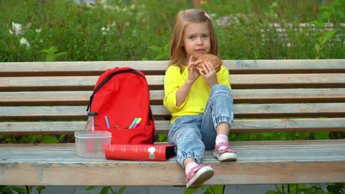 Child Sits on Bench with Backpack in Schoolyard and Eats Hamburger Education