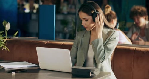Calm Attractive Young Woman in Buiisness Suit Wearing Headphones Earbuds Sits at Table at Cafe in