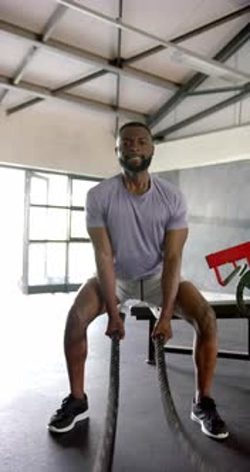 Vertical video: Man exercising with battle ropes in gym, smiling and focused on workout