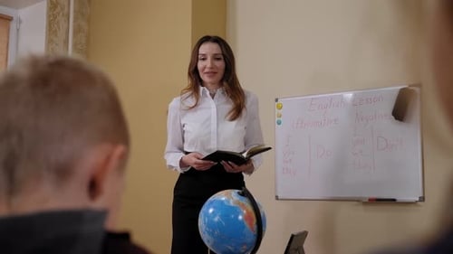A Smiling Teacher Stands at the Blackboard and Talks to the Children at School