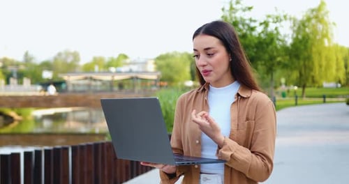 Young female freelancer working remotely in a park, focused on a video call using laptop. Outdoor