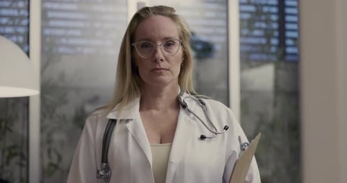 Serious female doctor holds clipboard and looks at camera in professional medical office with large