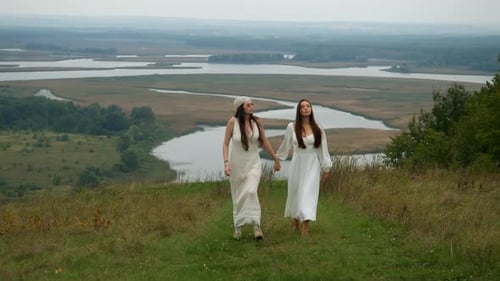 Young Women Walk Side By Side in Long White Dresses Surrounded By Wild Grass and Expansive River