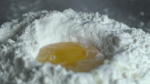 Whole raw egg falls into a pile of white flour then the chef's hand mixing the mix. Dough kneading e