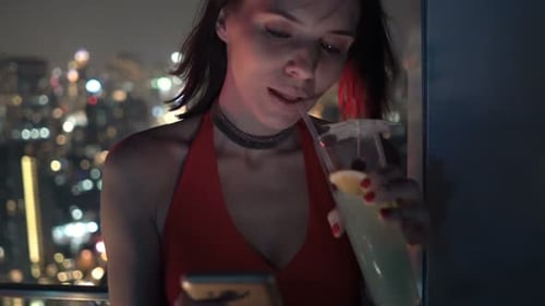 Elegant woman texting on smartphone and drinking cocktail at rooftop bar at night